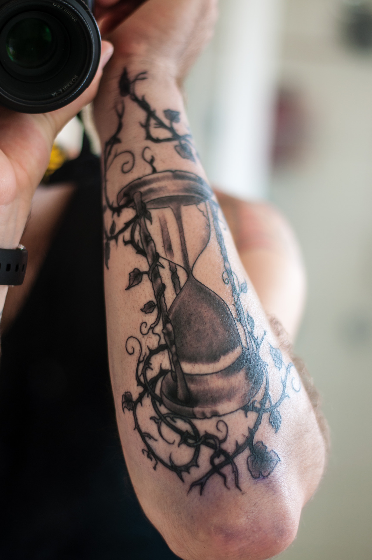 Black And Grey Hourglass With Vine Leaves Tattoo On Left Forearm