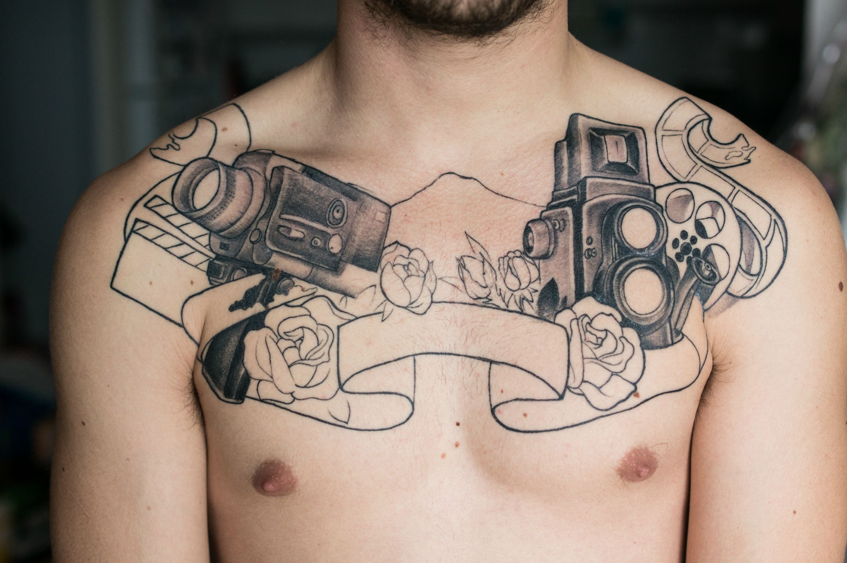 Two Movie Camera With Ribbon And Roses Tattoo On Man Chest.