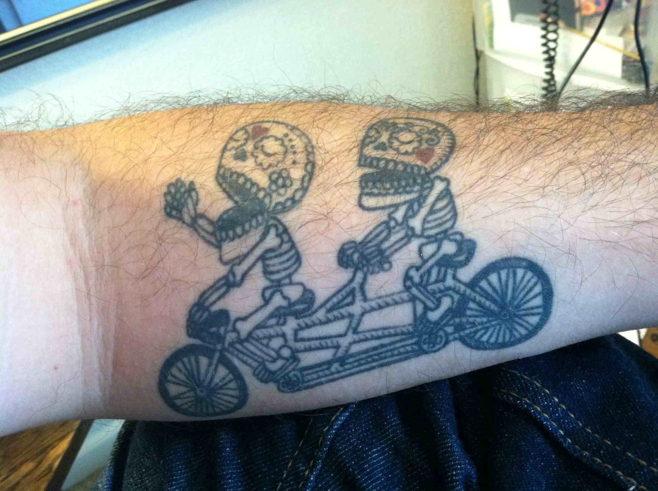 Two Sugar Skull Skeleton Riding Tandem Bike Tattoo On Forearm
