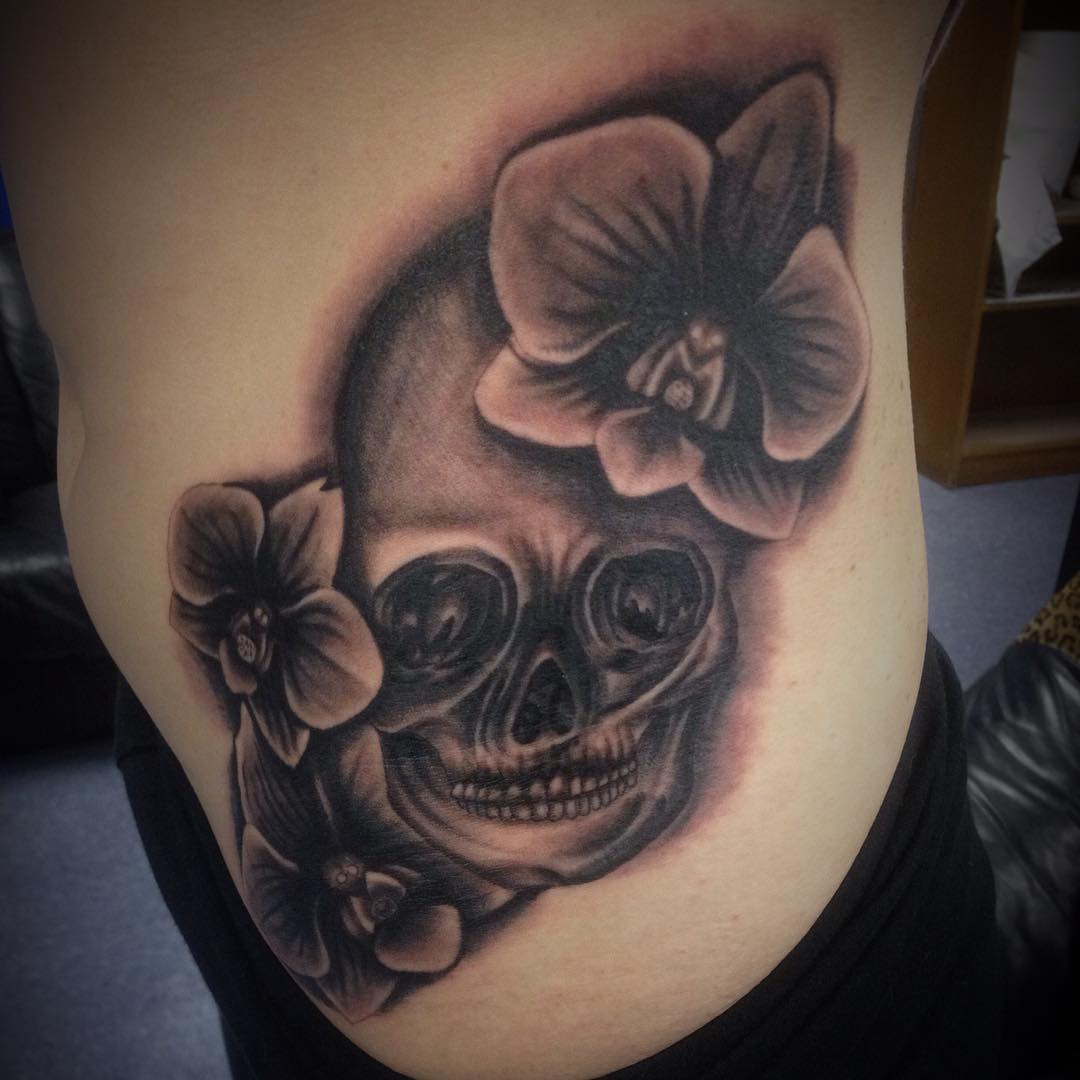 Realistic Grey Ink Skull And Orchid Tattoos On Side Rib