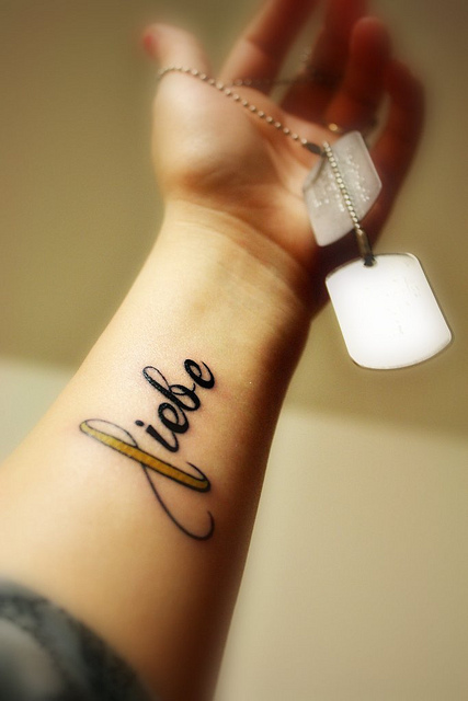 Liebe Word And Yellow Ribbon Tattoo On Left Forearm