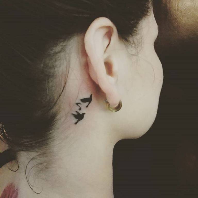 Black Flying Bird With Music Knot Tattoo On Right Behind The Ear