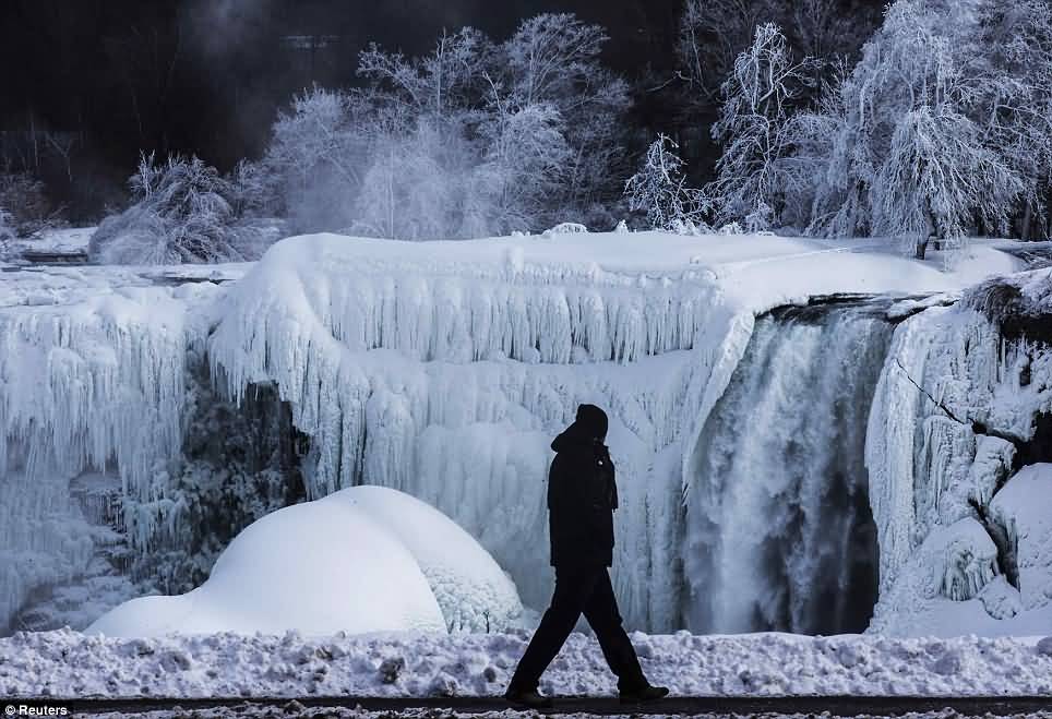 A Man Is Pictured Walking In Front Of Partially Frozen Niagara Falls From American Side