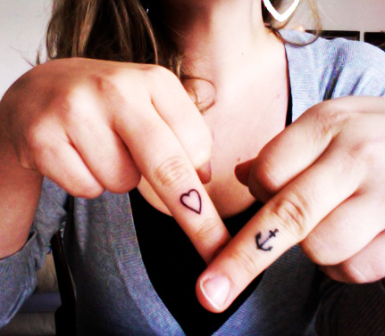 Girl Showing Her Anchor And Heart Tattoos On Fingers