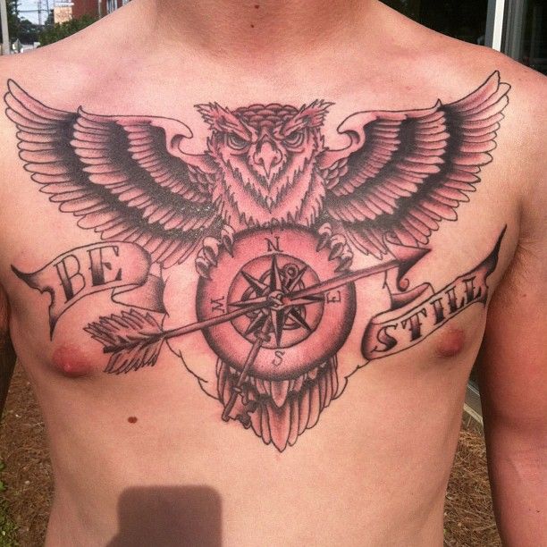 Black Ink Arrow In Compass With Flying Owl And Banner Tattoo On Man Chest