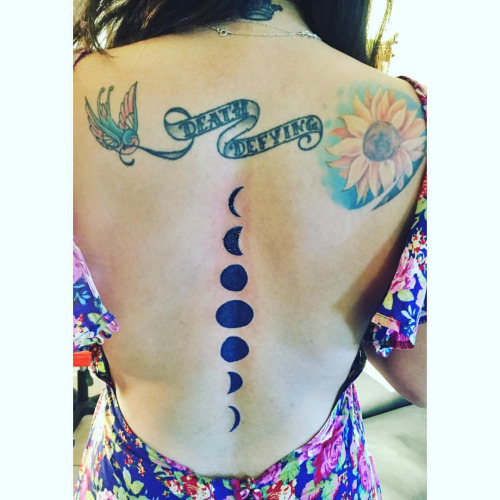 Death Defying Banner In Flying Bird Beak With Phases Of The Moon Tattoo On Girl Spine