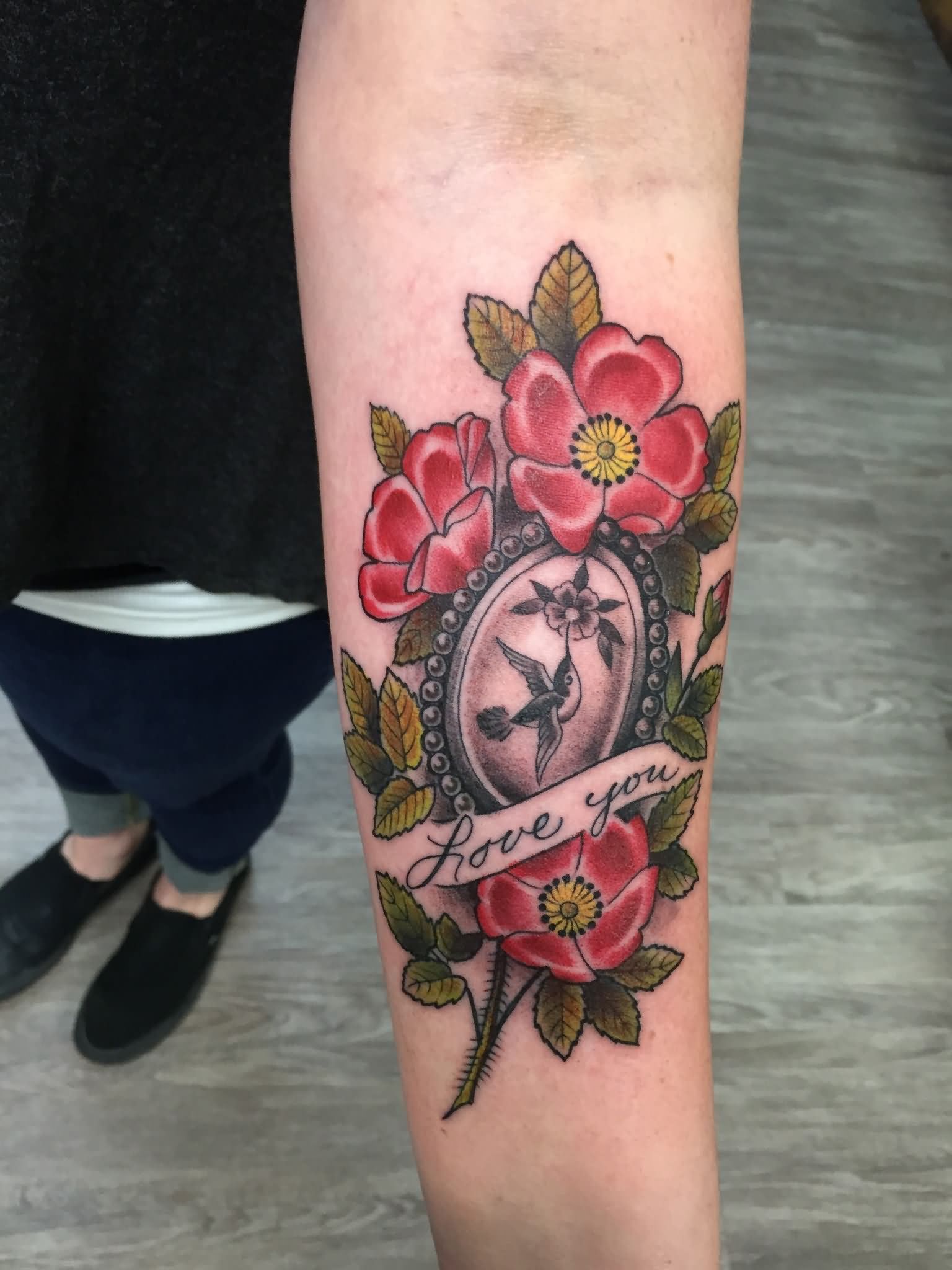Black Ink Bird In Frame With Flowers And Banner Tattoo On Left Forearm