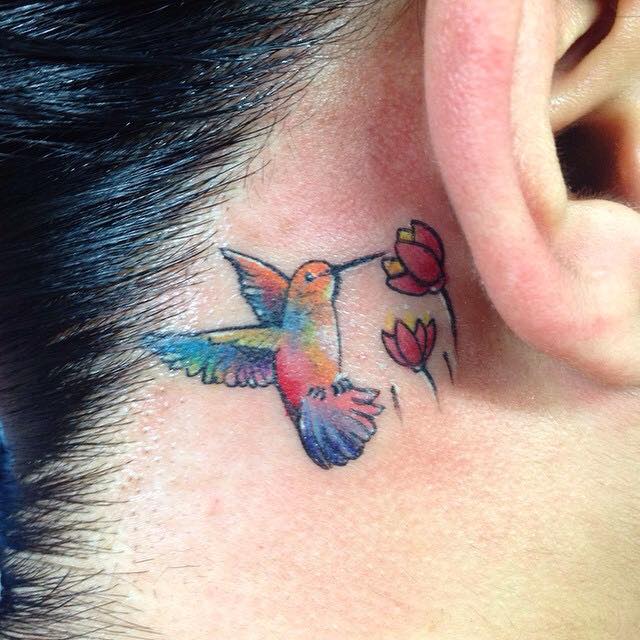 Colorful Flying Bird With Flowers Tattoo On Women Right Behind The Ear By Pig Legion