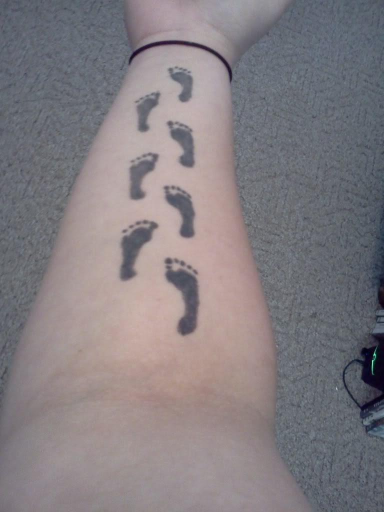 Black Ink Feet Prints Tattoo On Forearm