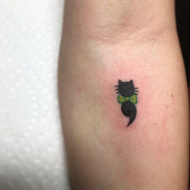 Beautiful Black Ink Semicolon Cat With Green Bow Tattoo On Forearm.