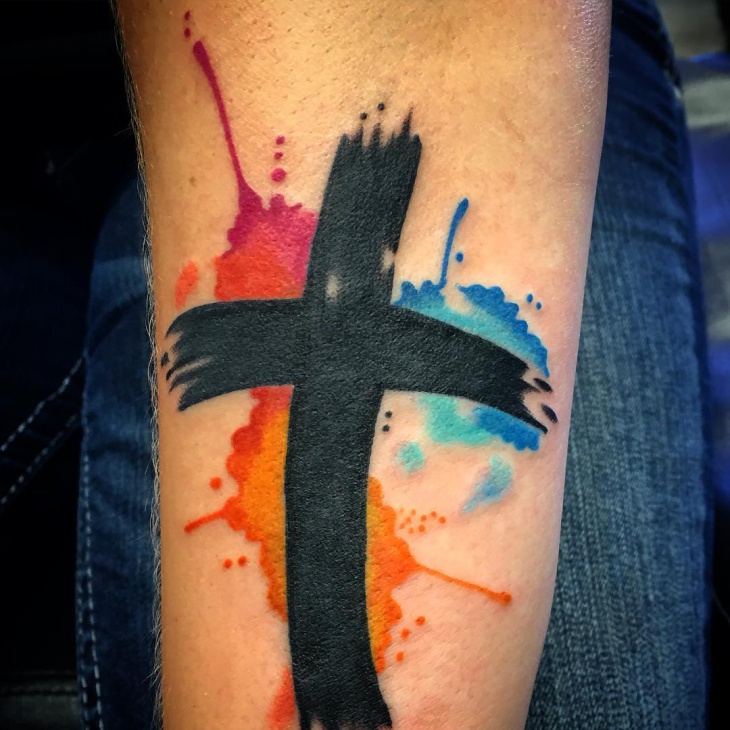 Black and colored cross with watercolor background tattoo on arm