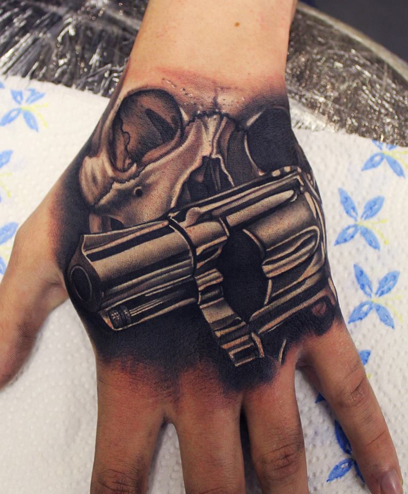 Grey shaded gun and skull tattoo on upper hand